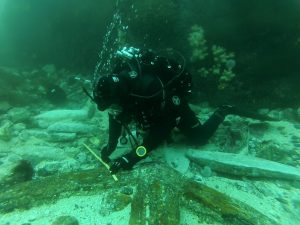 a diver in full SCUBA gear measuring a portion of shipwreck underwater