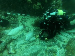 a diver in full SCUBA gear measuring a portion of a shipwreck underwater