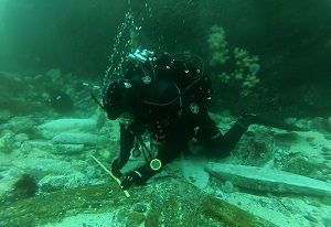 A diver in full SCUBA gear underwater measuring a portion of shipwreck (mark james)