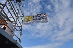 an image of the MSDS Marine flag waving in the wind. it is attached to a boat and behind the flag the sky is blue with small white clouds.