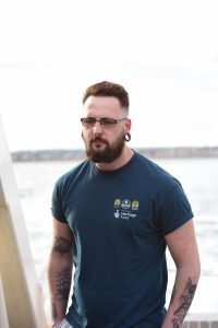 a man wearing a blue shirt with the MSDS Marine logo, heritage fund logo and go dive logo. he is wearing glasses. in the background there is out of focus water.