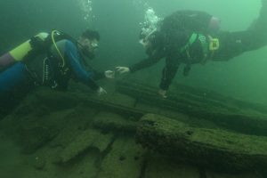 two divers in wetsuits with oxygen tanks are facing each other underwater. below them is the wood of a shipwreck.