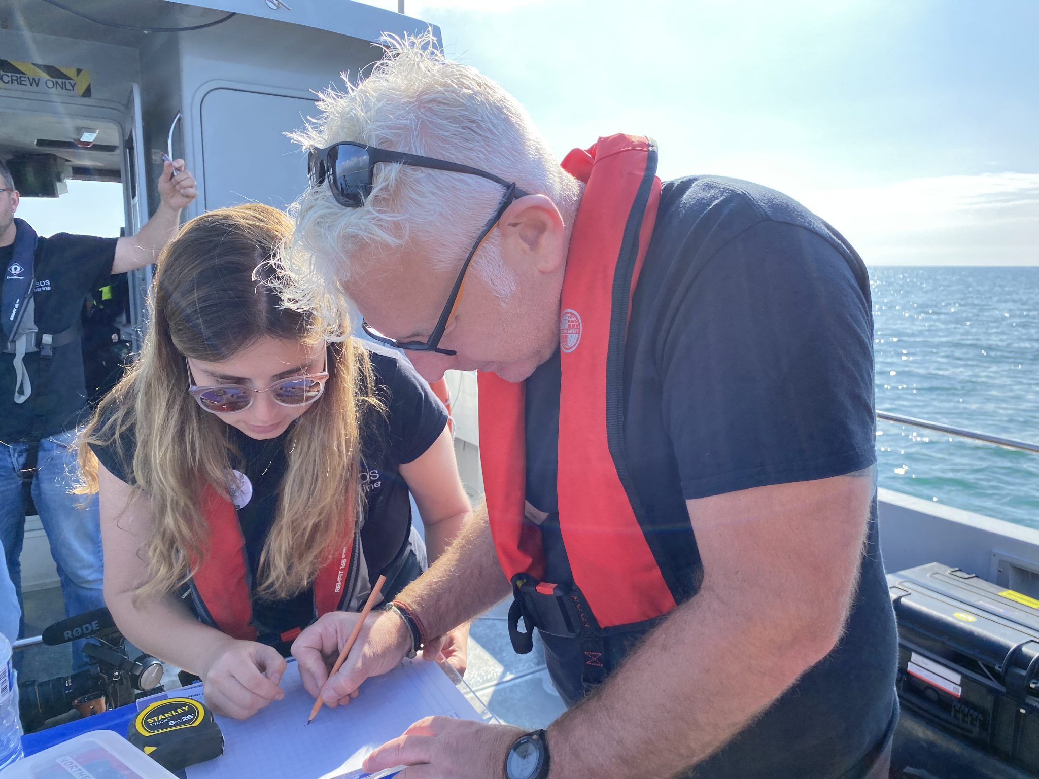 two people on a boat wearing life jackets leaning over a table. on the table is graph paper and a tape measure.