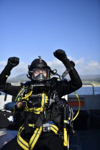 a diver wearing full SCUBA gear is sitting on a boat and doing a celebratory pose with both of their arms up.