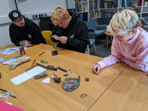 three people are sat at a table. on the table are several small objects. these objects are artefacts found on wrecks. two of the people are holding and inspecting one of the objects.