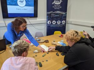 four people are seated around a table. on the table are a mix of small objects from wrecks. behind them is a poster for the nautical archaeological society. a woman (Peta Knott) in a blue shirt is pointing at one of the objects.