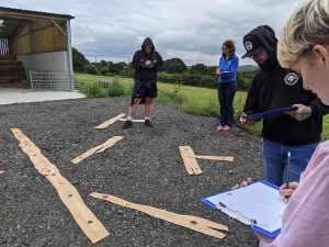 four people standing outside holding clipboards. there are seven fake pieces of wood on the floor in order to practice sketching wrecks.