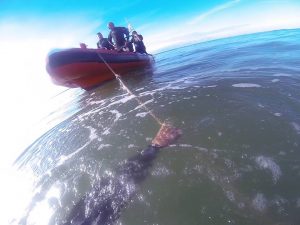 the arm of a diver below the water holding onto a rope attached to an orange boat with several other divers standing in it