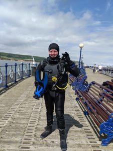 a diver (James MacDonell) standing on a pier in a wetsuit and holding hand up to give the 'ok' symbol.